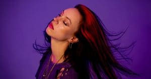 Female music artist with flowing red hair and closed eyes against purple backdrop.