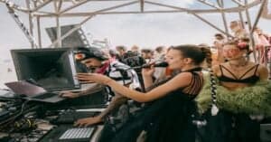 DJ mixing music at outdoor festival with crowd and sky in background.